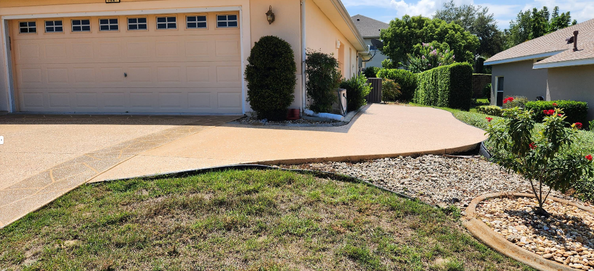 Tan painted concrete driveway leading to beige home with manicured shrubs and decorative rock landscaping