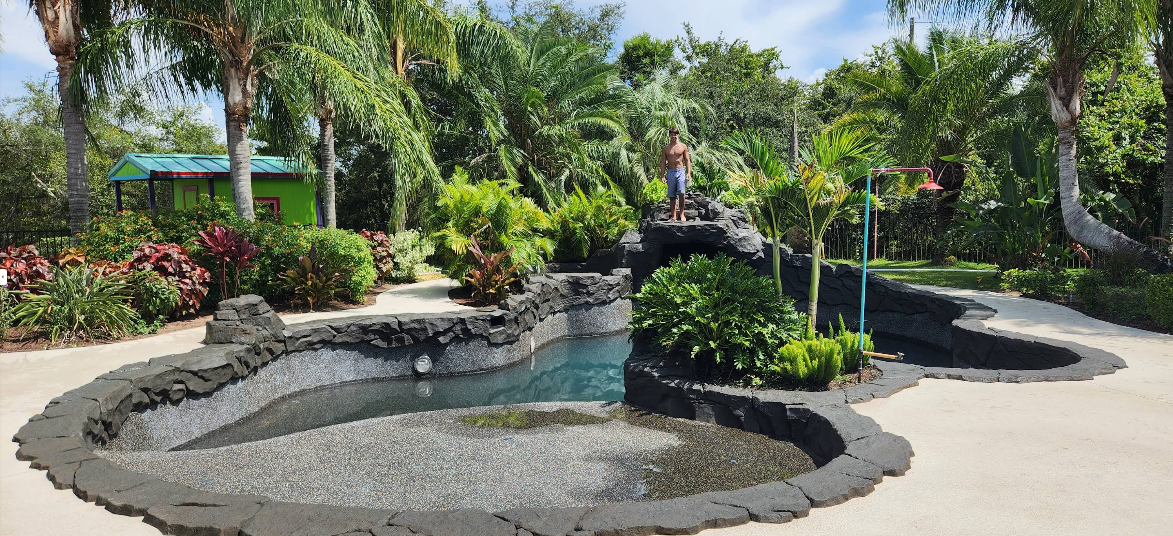 Custom pool with rock waterfall feature and light painted concrete deck surrounded by tropical landscaping