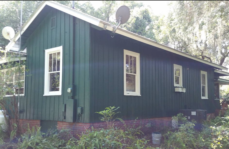 Small home exterior painted in dark teal green with white trim windows surrounded by greenery