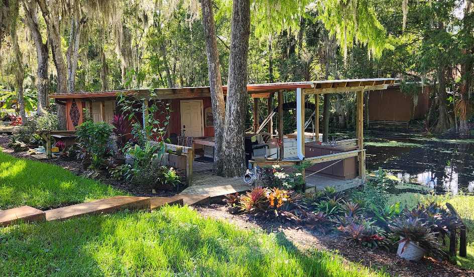 Backyard view of painted outbuilding structures with wooden decking under moss-draped trees and natural landscaping