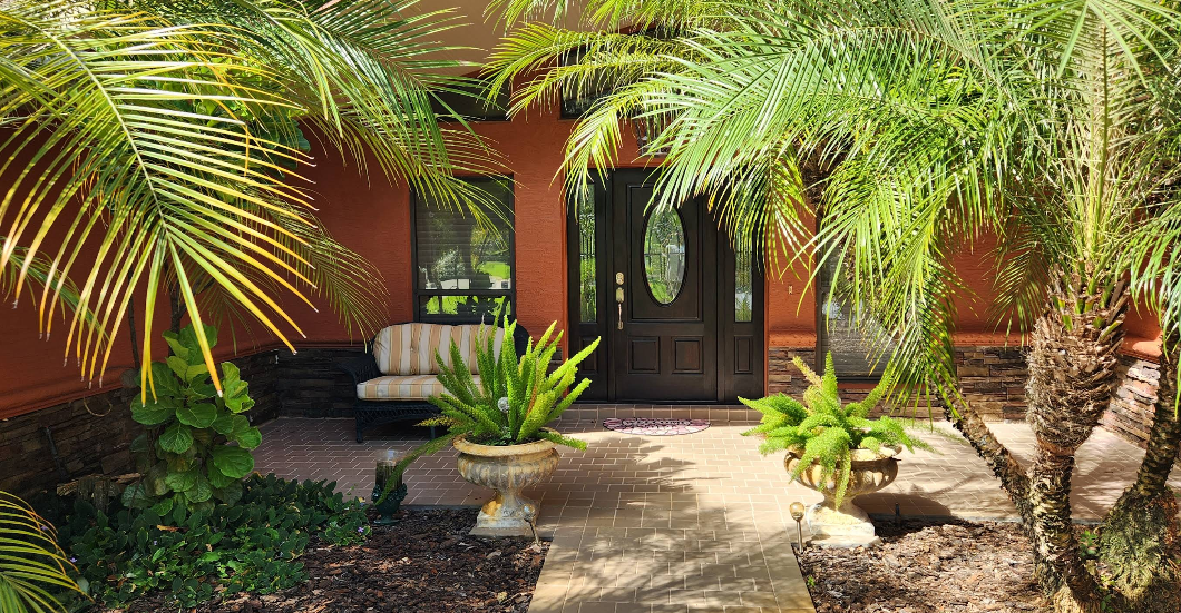 Home entrance painted in rust orange with dark door, brick paver walkway and tropical landscaping with palm trees
