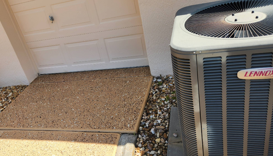 Tan speckled painted concrete pad beside white garage door and HVAC unit with pebble landscaping