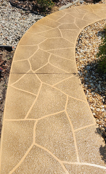Curved tan painted concrete walkway with flagstone pattern through pebble rock landscaping