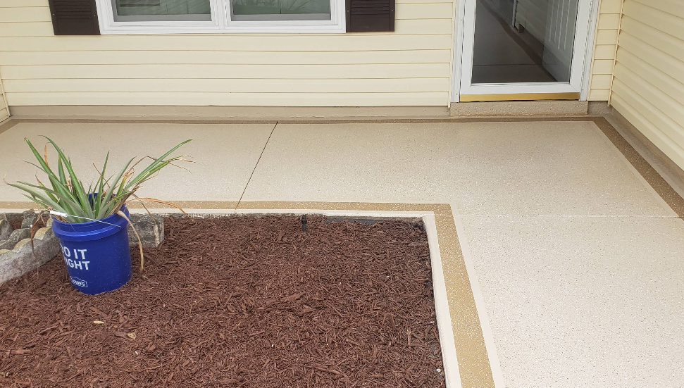 Beige painted concrete patio with tan decorative border and mulched planting area at yellow home