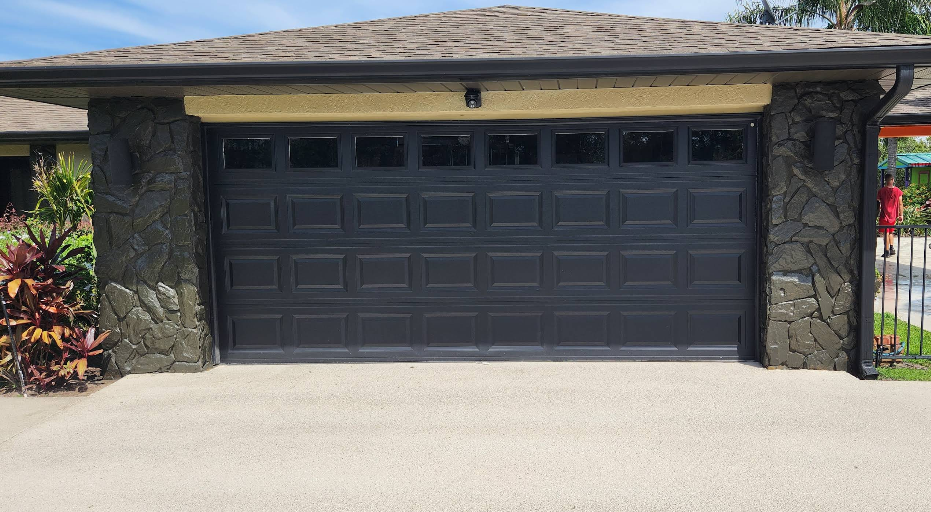 Light beige painted concrete driveway in front of home with dark painted garage door and stone pillar accents