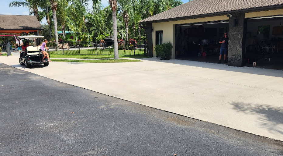 Wide light beige painted concrete driveway with golf cart and open garage showing tropical landscaping